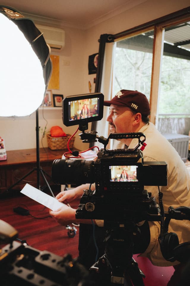 A camera operator films a woman in a lounge room, with monitors displaying the live shot on set.