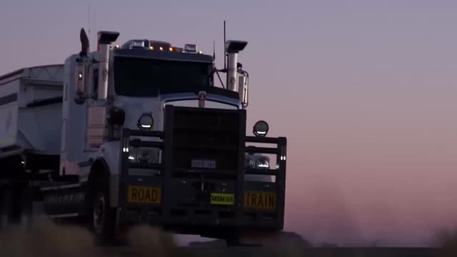 A road train truck marked 'Road Train' drives along a rural road at dusk.