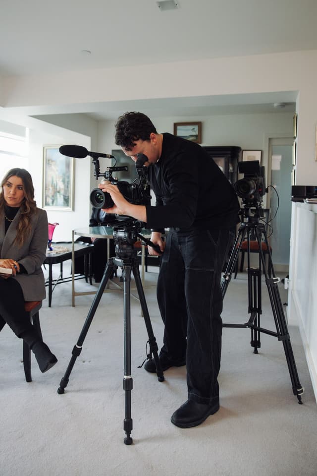 A videographer films with a cinema camera on a tripod while a woman sits in the background.