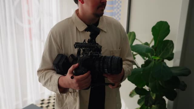 A man holds a cinema camera rig with a large lens beside an indoor fig plant.