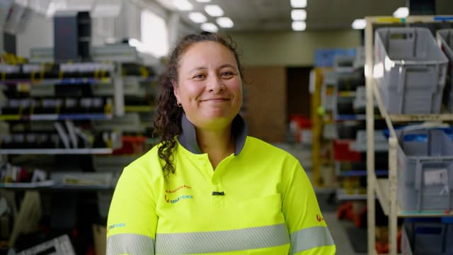 An Australia Post StarTrack worker in a yellow hi-vis shirt smiles inside a sorting warehouse.