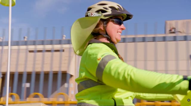 An Australia Post employee in high-vis and a helmet smiles while cycling through a depot at 11:20am.