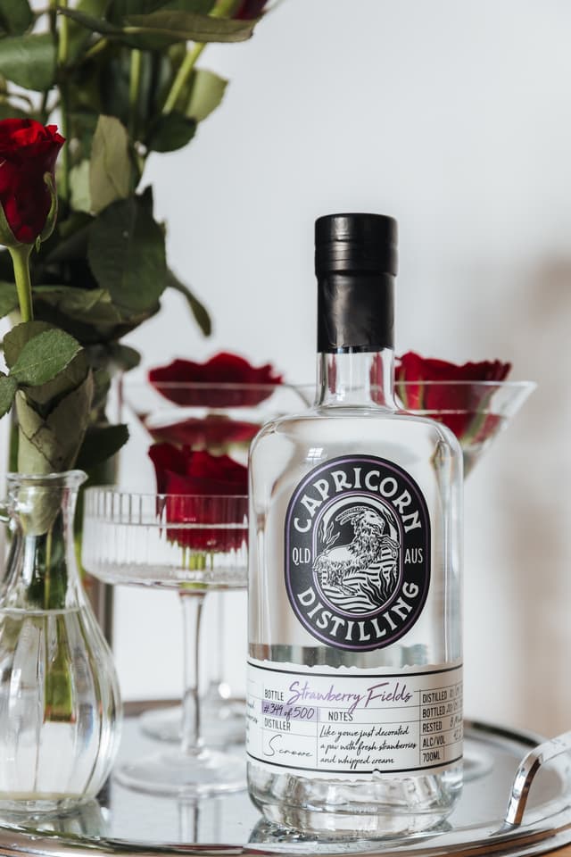 A Capricorn Distilling gin bottle labelled Strawberry Fields displayed beside a vase of red roses.
