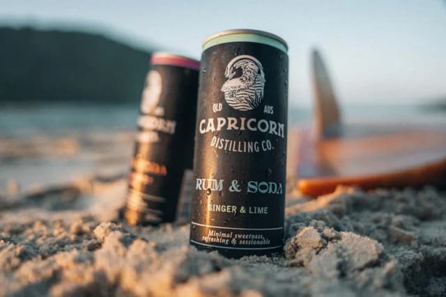 Two Capricorn Distilling Co rum and soda cans stand in beach sand with a surfboard blurred behind them.