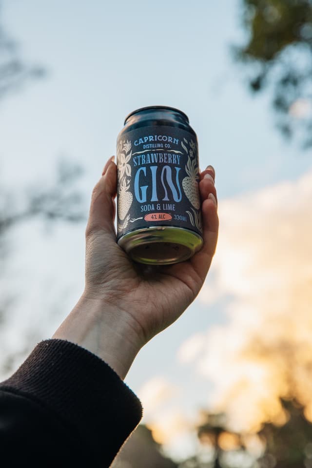 A hand raises a can of Capricorn Distilling Co Strawberry Gin soda and lime against a sunset sky.