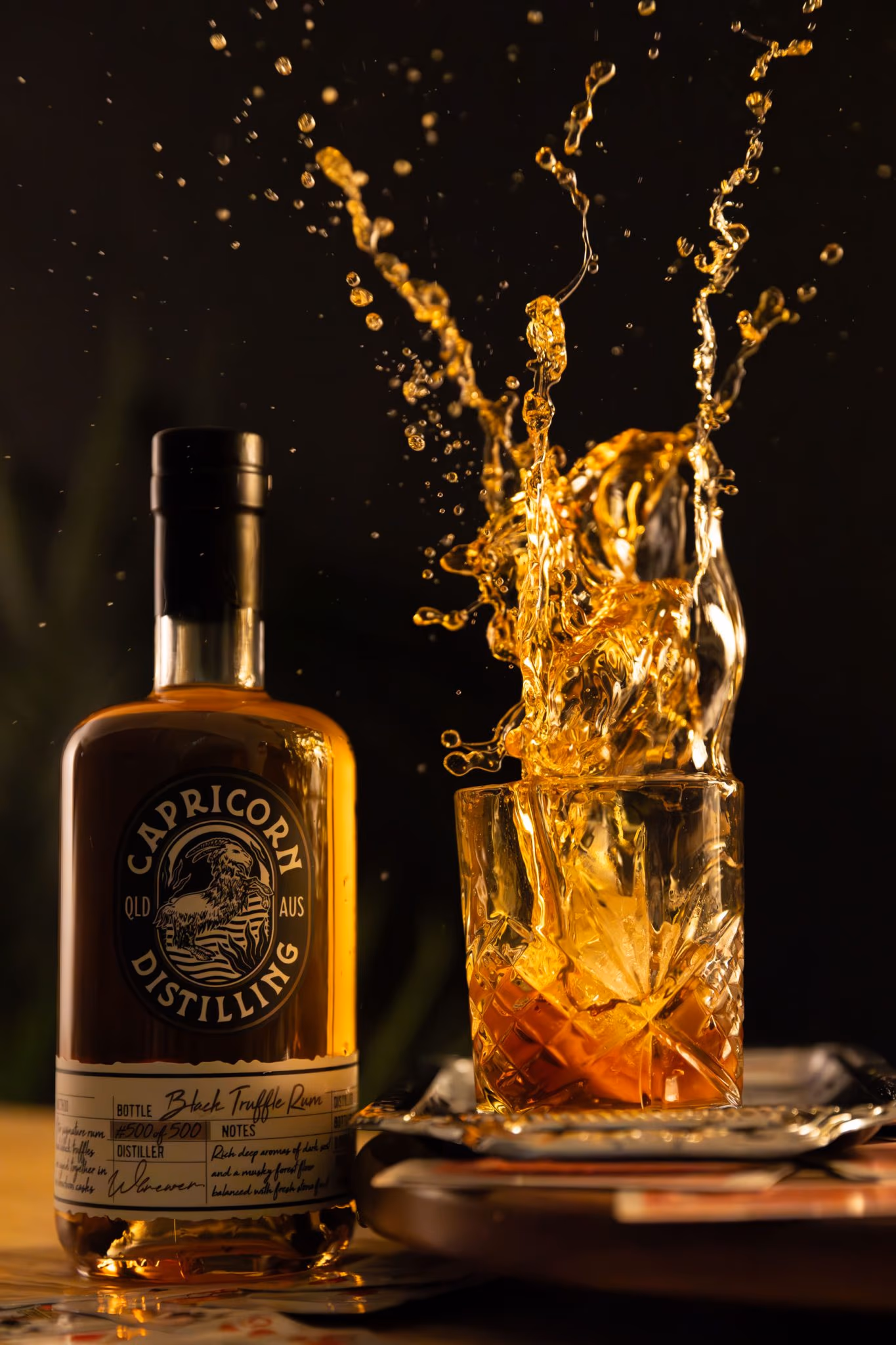 A Capricorn Distilling Co Black Truffle Rum bottle beside a crystal tumbler with rum splashing up out of it.