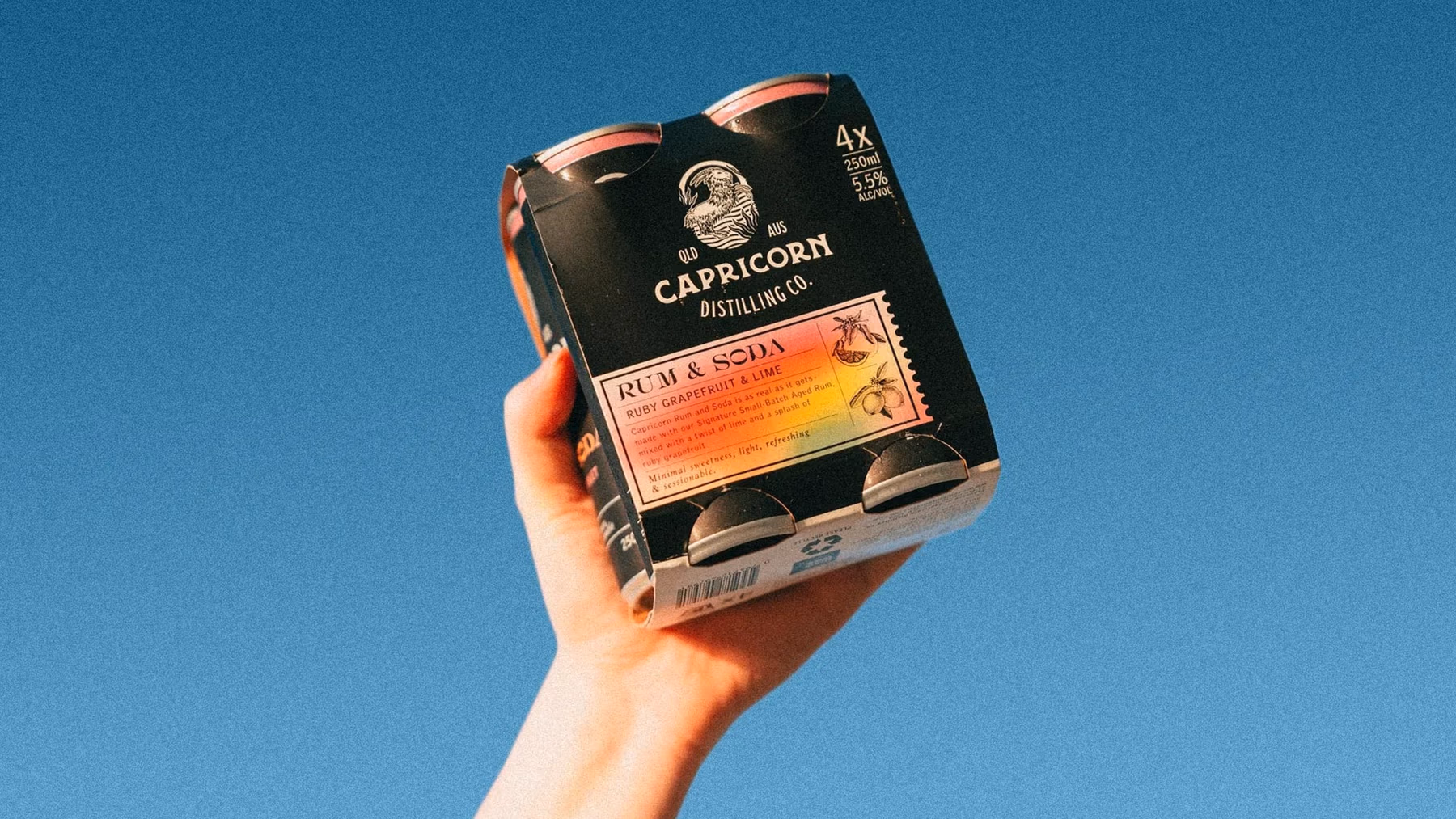 A hand holds a four pack of Capricorn Distilling Co Rum and Soda cans up against a clear blue sky.