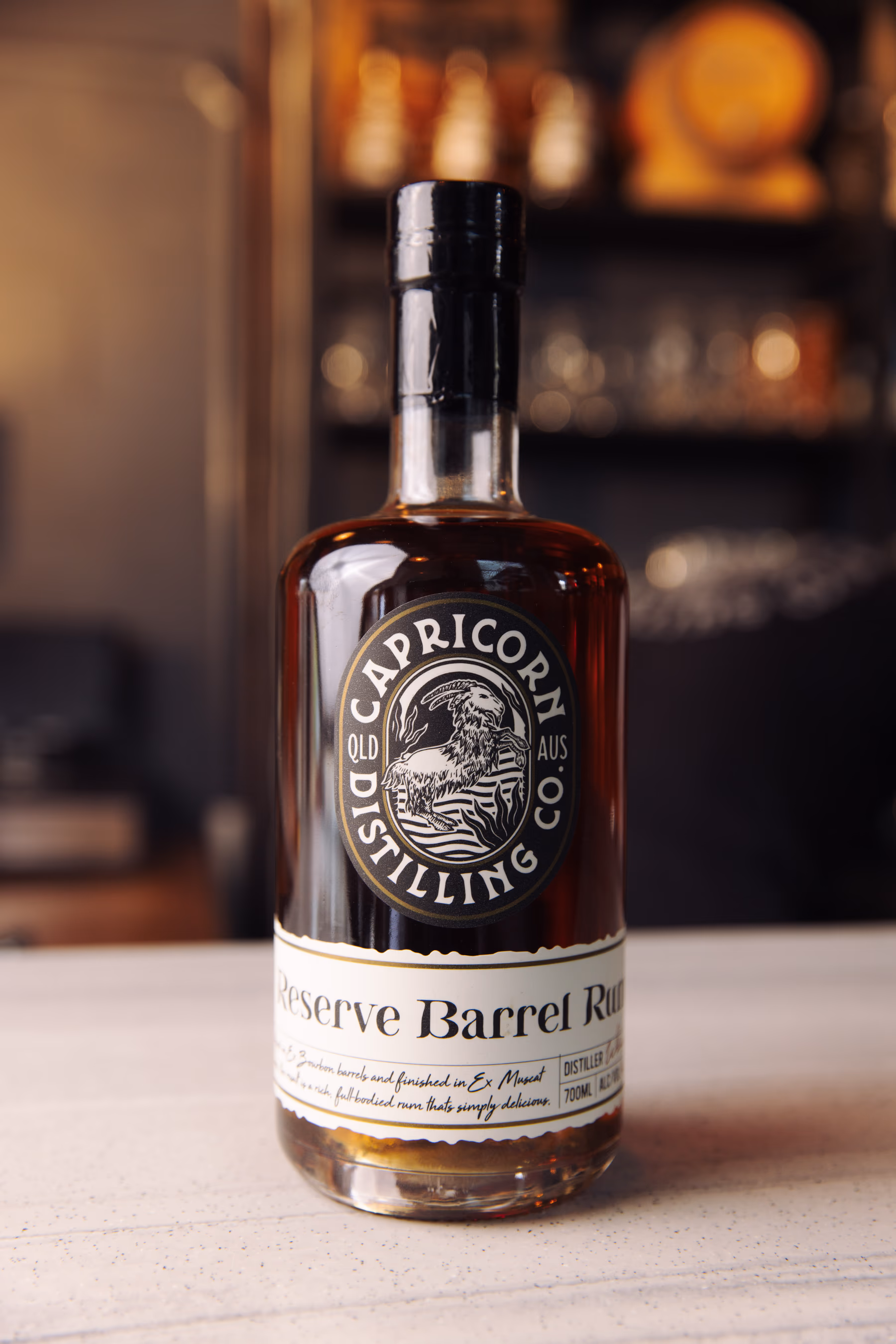 A bottle of Capricorn Distilling Co Reserve Barrel Rum stands on a bar counter with barrels blurred behind it.