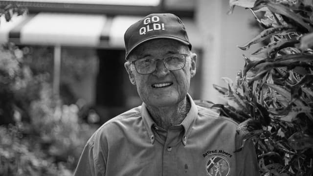 A black and white portrait of a smiling elderly man wearing a GO QLD cap, for Heart of Australia.