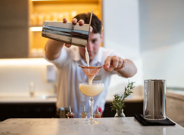 A bartender pours a cocktail through a strainer into a coupe glass beside a rosemary sprig at Hilton Surfers Paradise.