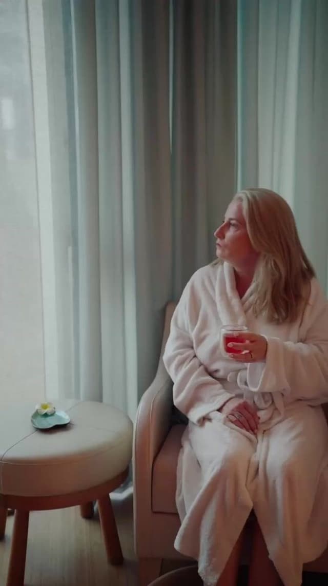 A woman in a white robe sits by a curtained window with a drink at Hilton Surfers Paradise.