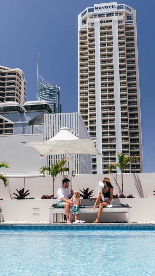 A family sits poolside at Hilton Surfers Paradise with Gold Coast high-rises, including the Novotel tower, behind them.