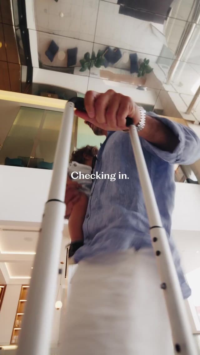 A low angle shot of a man pushing a luggage cart with a child nearby, captioned 'Checking in' at Hilton Surfers Paradise.