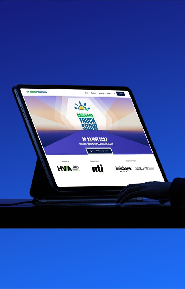 A hand holds a tablet displaying the Heavy Vehicle Industry Australia Brisbane Truck Show website.