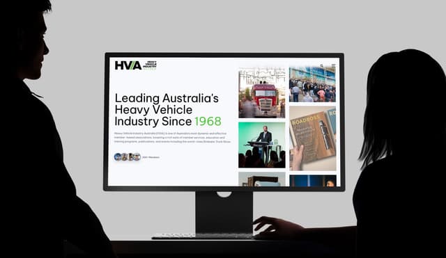 Two silhouetted figures view the Heavy Vehicle Industry Australia website homepage on a large monitor.