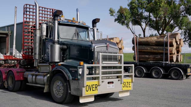 A Kenworth logging truck marked 'Over Size' hauls a load of logs past a shed and trees.