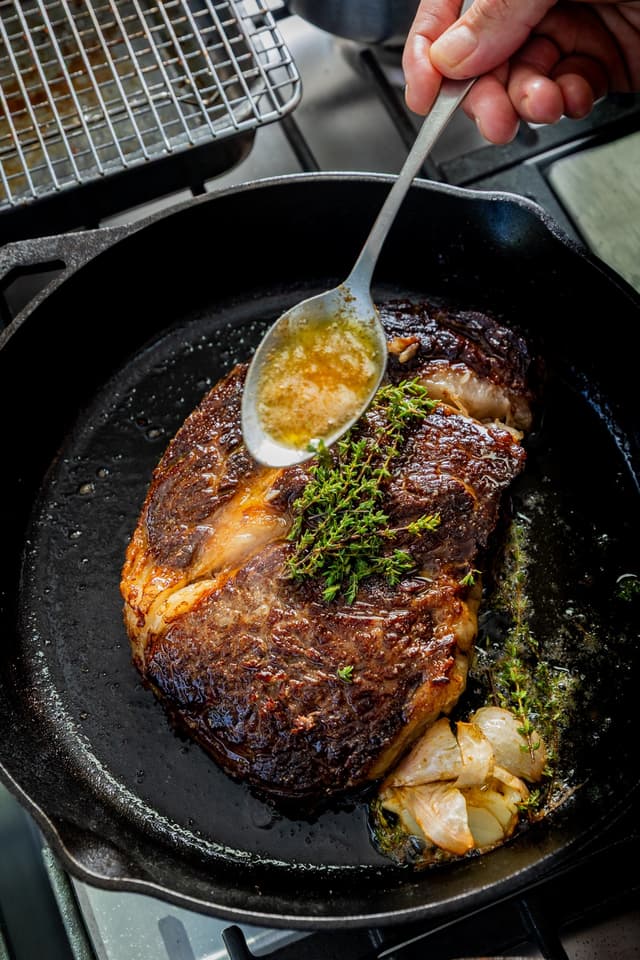 A hand spoons herb butter over a seared steak with garlic and thyme in a cast iron skillet, a Providore Global dish.