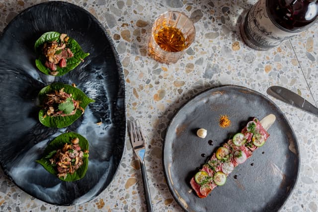 Betel leaf wraps and a beef tartare plate are served with a whisky cocktail on a terrazzo table, a Providore Global dish.
