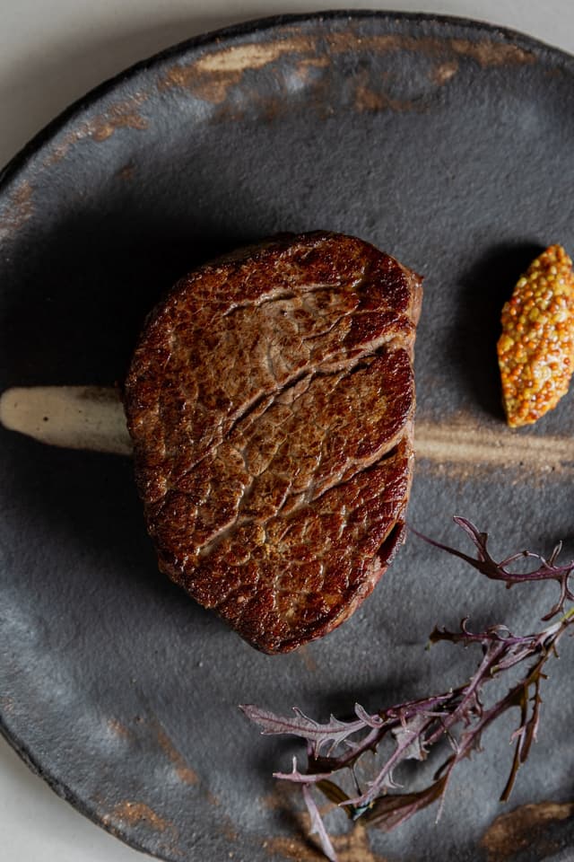 A seared eye fillet steak rests on a dark plate with wholegrain mustard and purple mustard greens, a Providore Global dish.