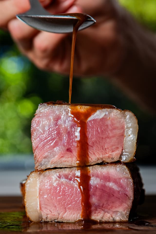 A hand drizzles dark sauce over two stacked slices of medium rare steak, a Providore Global dish.