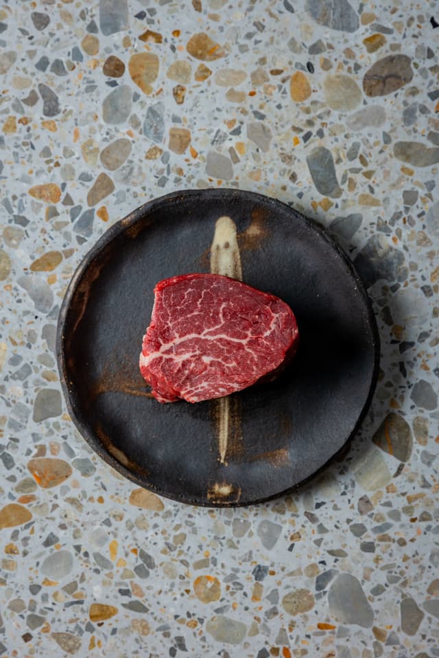 An overhead shot of a marbled raw steak on a dark ceramic plate atop a terrazzo surface, a Providore Global dish.