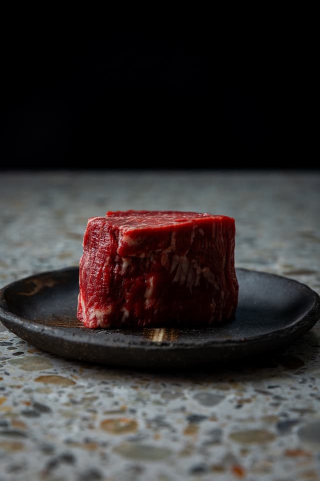 A raw beef fillet steak sits on a dark ceramic plate against a black background, from a Providore Global shoot.