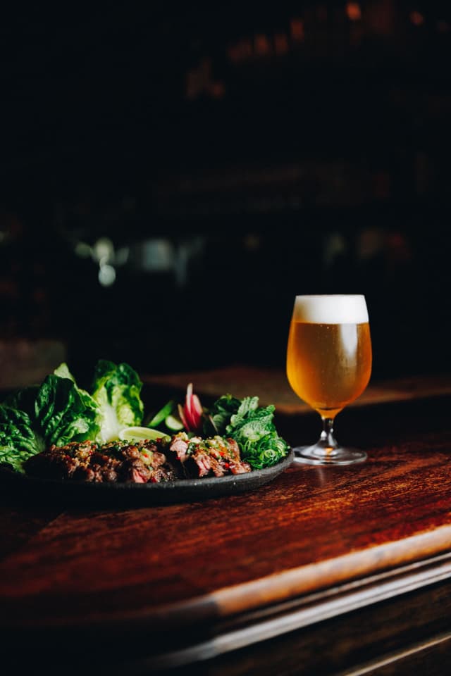 A plate of grilled sliced meat with salad greens beside a glass of beer on a dark wooden table at a Providore Global venue.