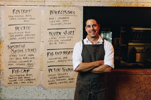 A Providore Global chef stands arms crossed in front of handwritten menus listing cuts like rib cap and deckle.
