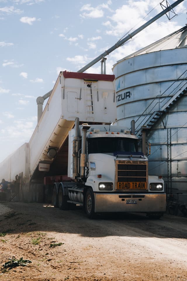 A Providore Global road train tips grain into a silo as dust rises under a clear blue sky.