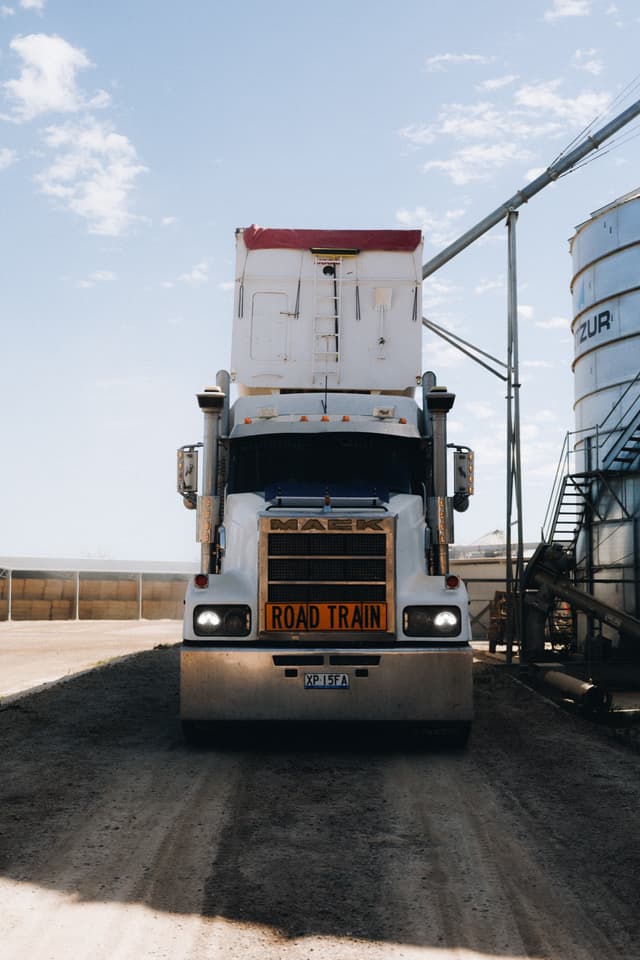 The front of a Providore Global Mack road train parked beside a grain silo with its headlights on.