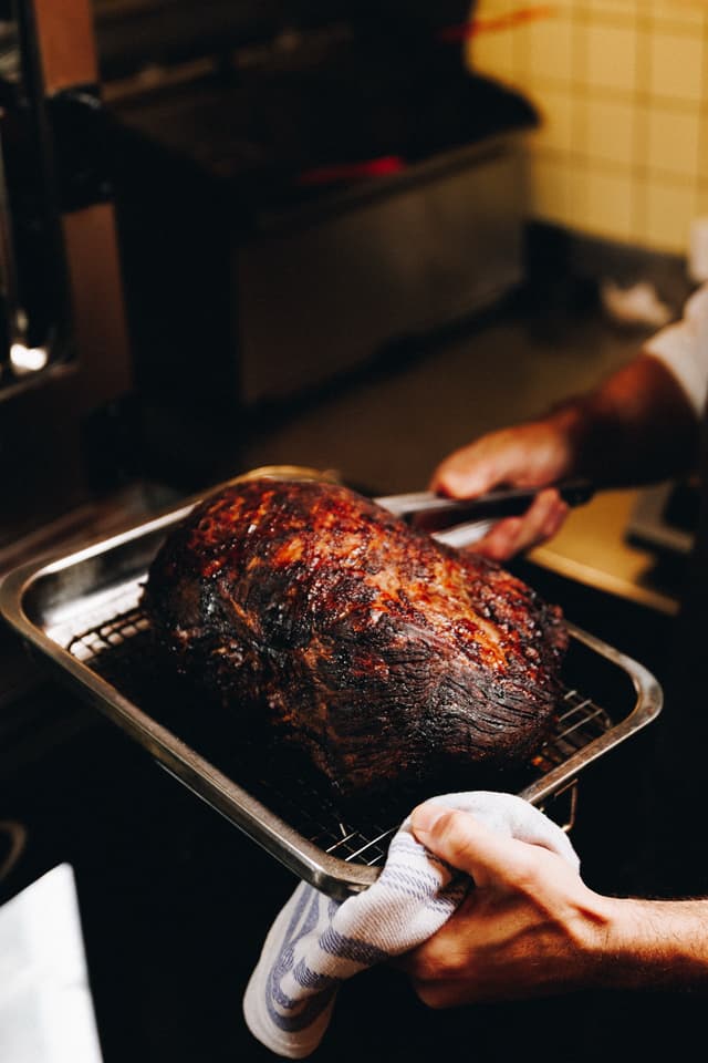 A chef lifts a large roasted cut of Providore Global meat from the oven on a tray with tongs.