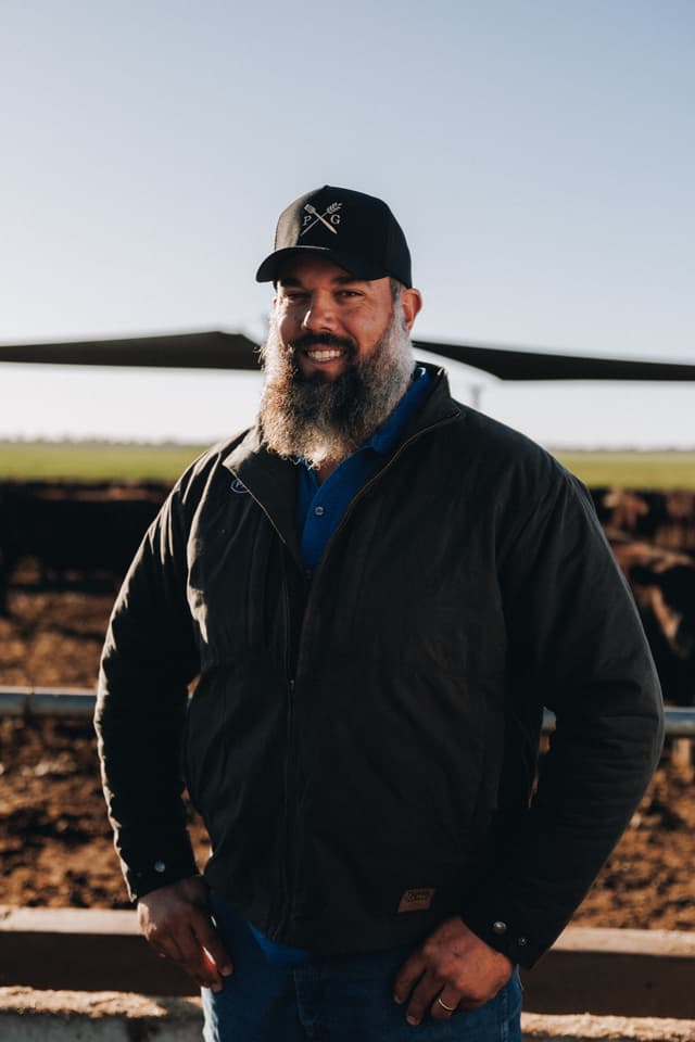 A bearded man in a Providore Global cap and jacket smiles in front of cattle at a feedlot.