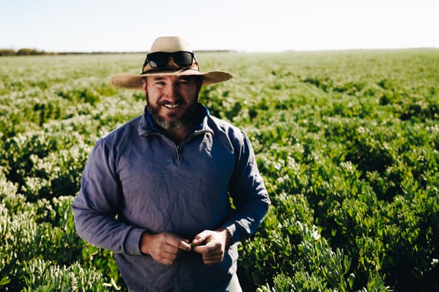 A Providore Global grower in a straw hat and sunglasses smiles while standing in a flowering green field.