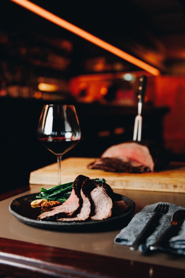Sliced roast beef with charred greens on a plate at a Providore Global venue, red wine glass and neon light overhead.
