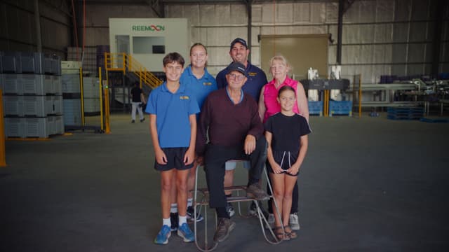 Three generations of a family pose together inside the Reemoon warehouse among stacked crates and processing machinery.