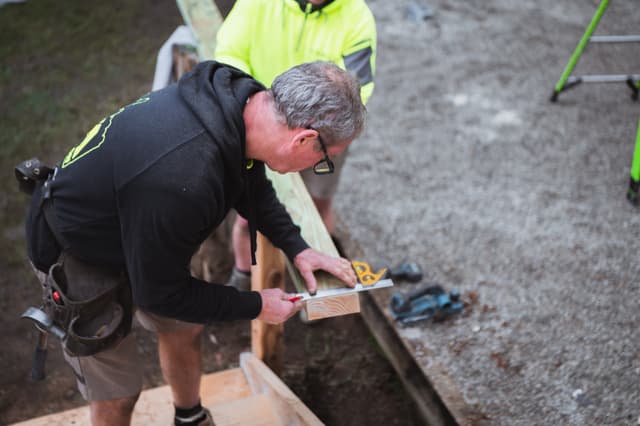 A tradesman marks a timber stud with a square while a colleague in hi-vis looks on.