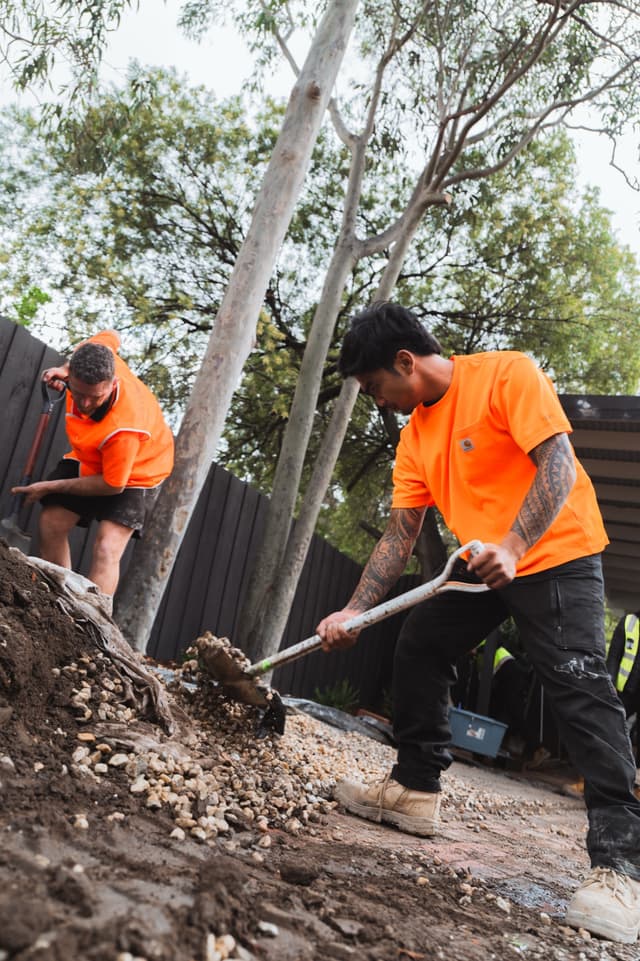 Two tradesmen in hi-vis shovel gravel and soil on a landscaped construction site.