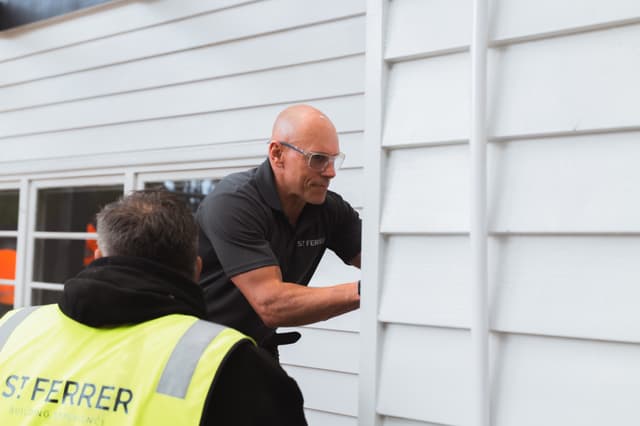 A St Ferrer builder in a branded polo fits weatherboard cladding on a house while a colleague in a St Ferrer hi-vis vest looks on.