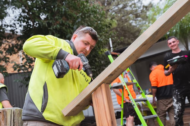 A construction worker in high vis drills a timber frame while colleagues work in the background of a building site.