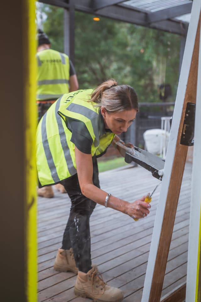 A tradeswoman in a yellow hi-vis vest paints a door frame on a timber deck, with a colleague working in the background.