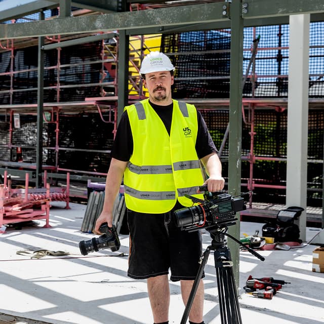 An Usher Group crew member in a hard hat and hi-vis vest holds camera equipment on a scaffolded construction site.
