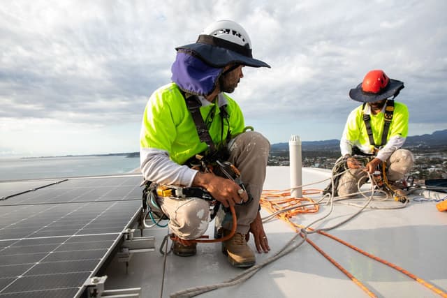 Two workers in high-vis gear install solar panels on a rooftop overlooking the coastline for Usher Group.