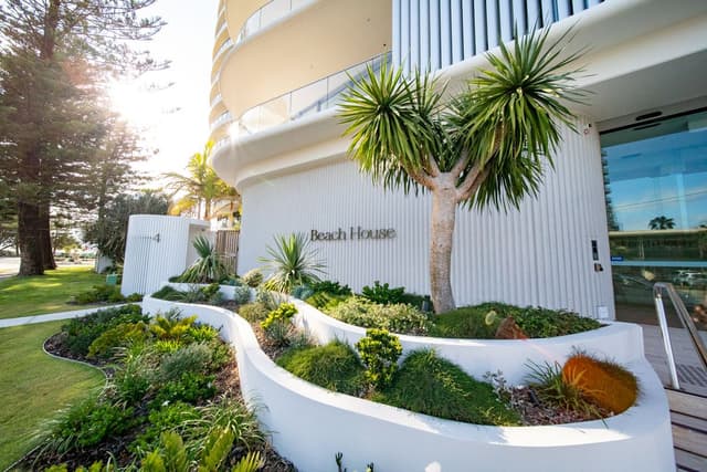 The Beach House development by Usher Group features a curved white facade, a palm tree, and landscaped garden beds.