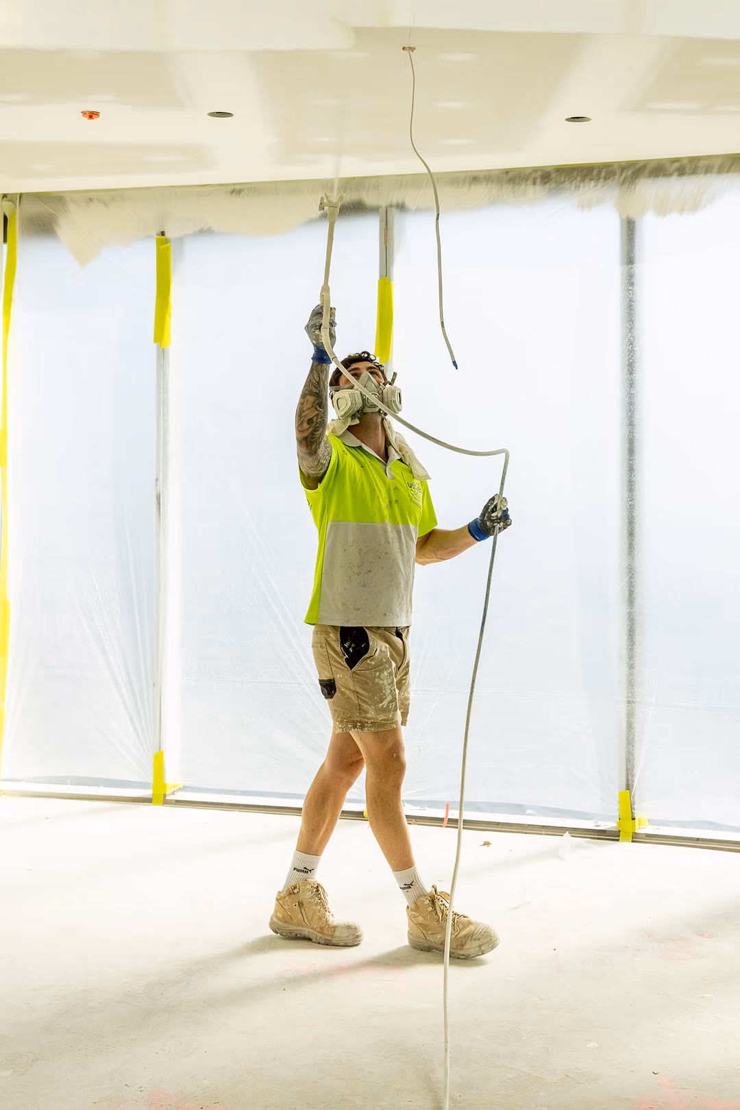An Usher Group worker in a respirator and hi-vis spray paints a ceiling inside a masked-up apartment.