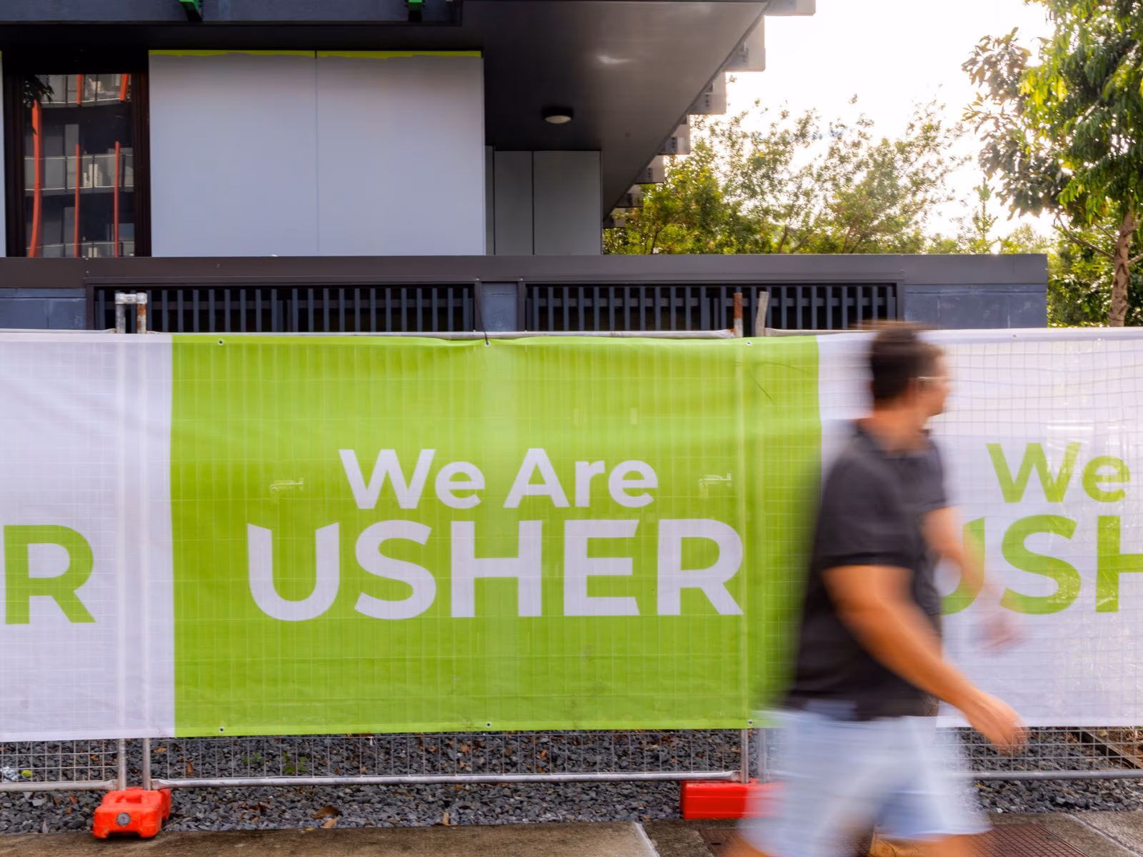 A worker walks past green Usher Group site hoarding reading We Are Usher outside a new building.