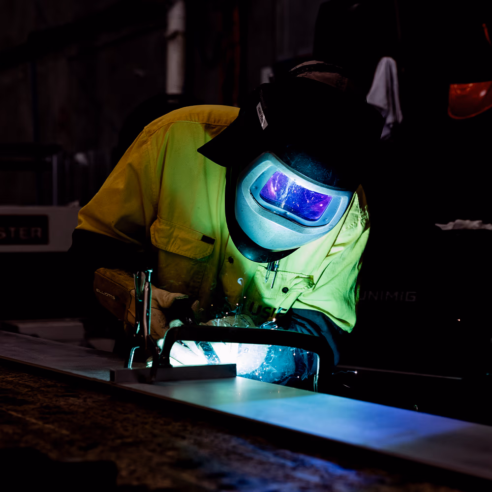 An Usher Group tradesperson in a welding helmet and hi-vis shirt welds steel as sparks light the workshop.