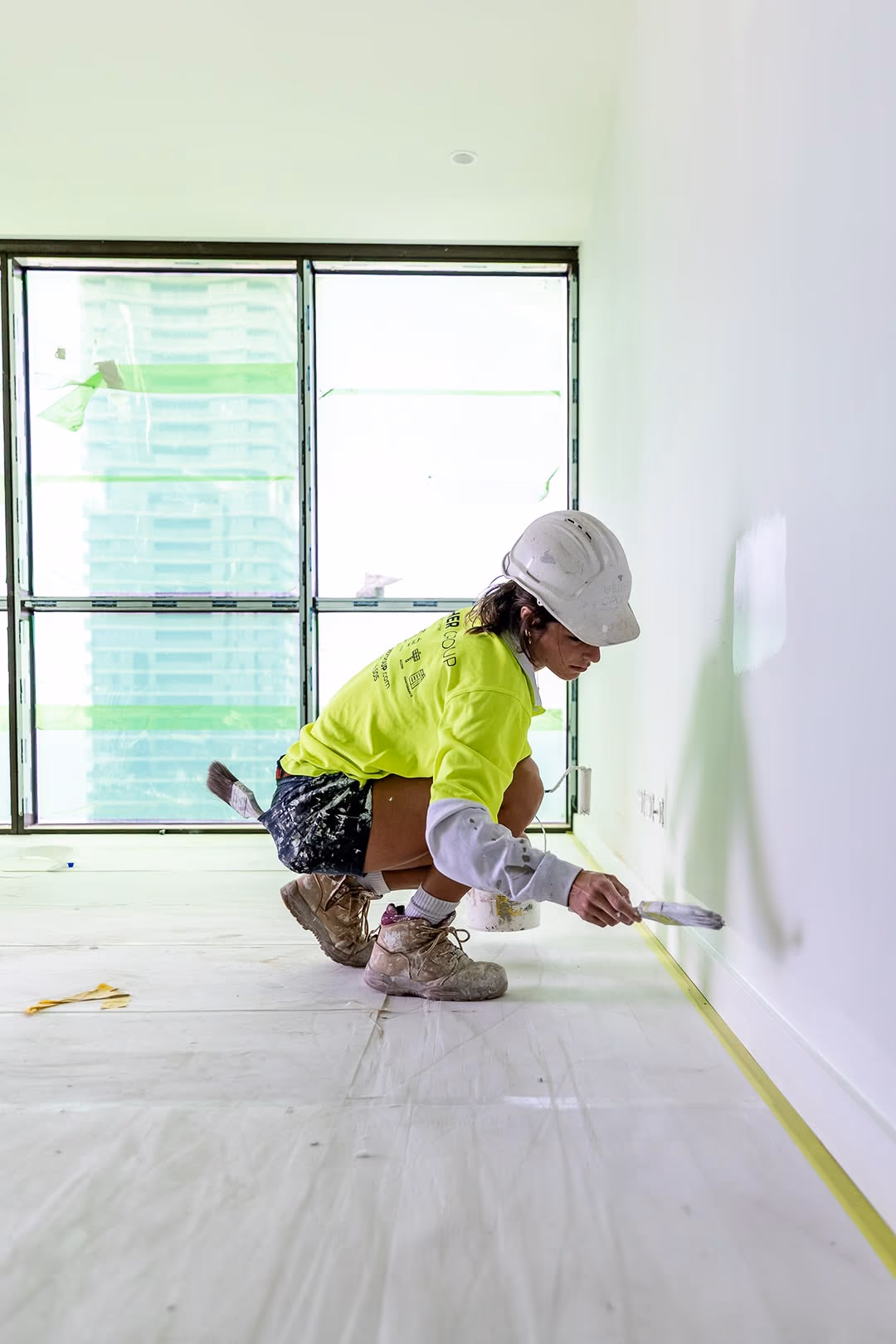 An Usher Group painter in a hard hat and hi-vis crouches to cut in paint along a skirting line.