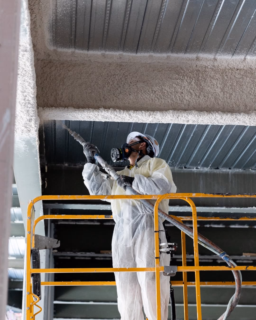 An Usher Group applicator in a white suit and respirator sprays protective coating onto steel from a scissor lift.