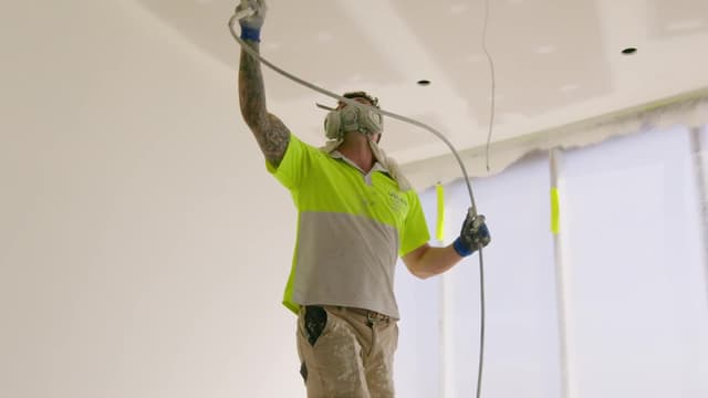 A tradesman in a hi-vis Usher Group shirt and respirator mask applies plaster to a ceiling with a spray hose.