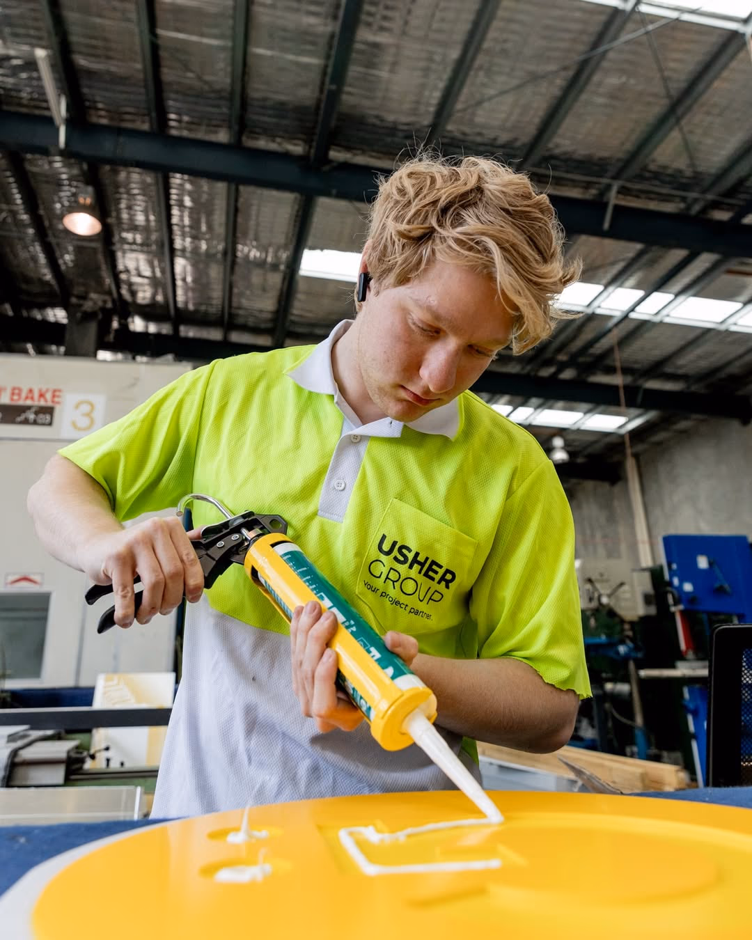 A young Usher Group worker in a hi-vis polo applies sealant to a yellow panel in a workshop.